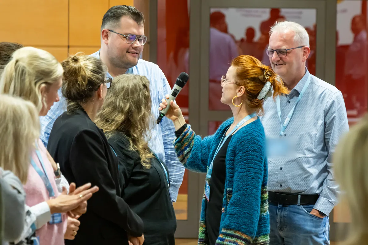 Christiane Schmieder mit Mikrofon im Gespräch mit einer Gruppe von Kommunalpolitikerinnen und Kommunalpolitikern